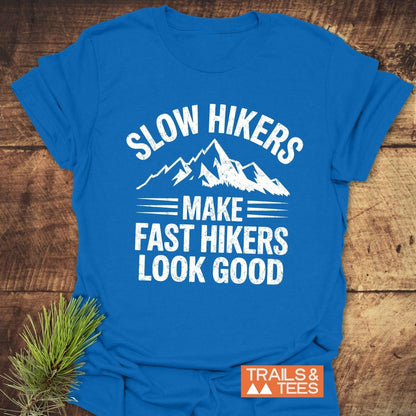 Royal-blue Slow Hikers T-Shirt with mountain graphic and slogan, featuring TRAILS & TEES logo, laid on wooden surface with pine sprig.