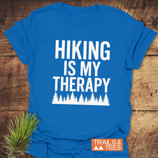 Hiking Is My Therapy T-Shirt on wooden surface, featuring bold white text and pine tree silhouette, styled with pine needles; made from soft, durable cotton by Trails & Tees.