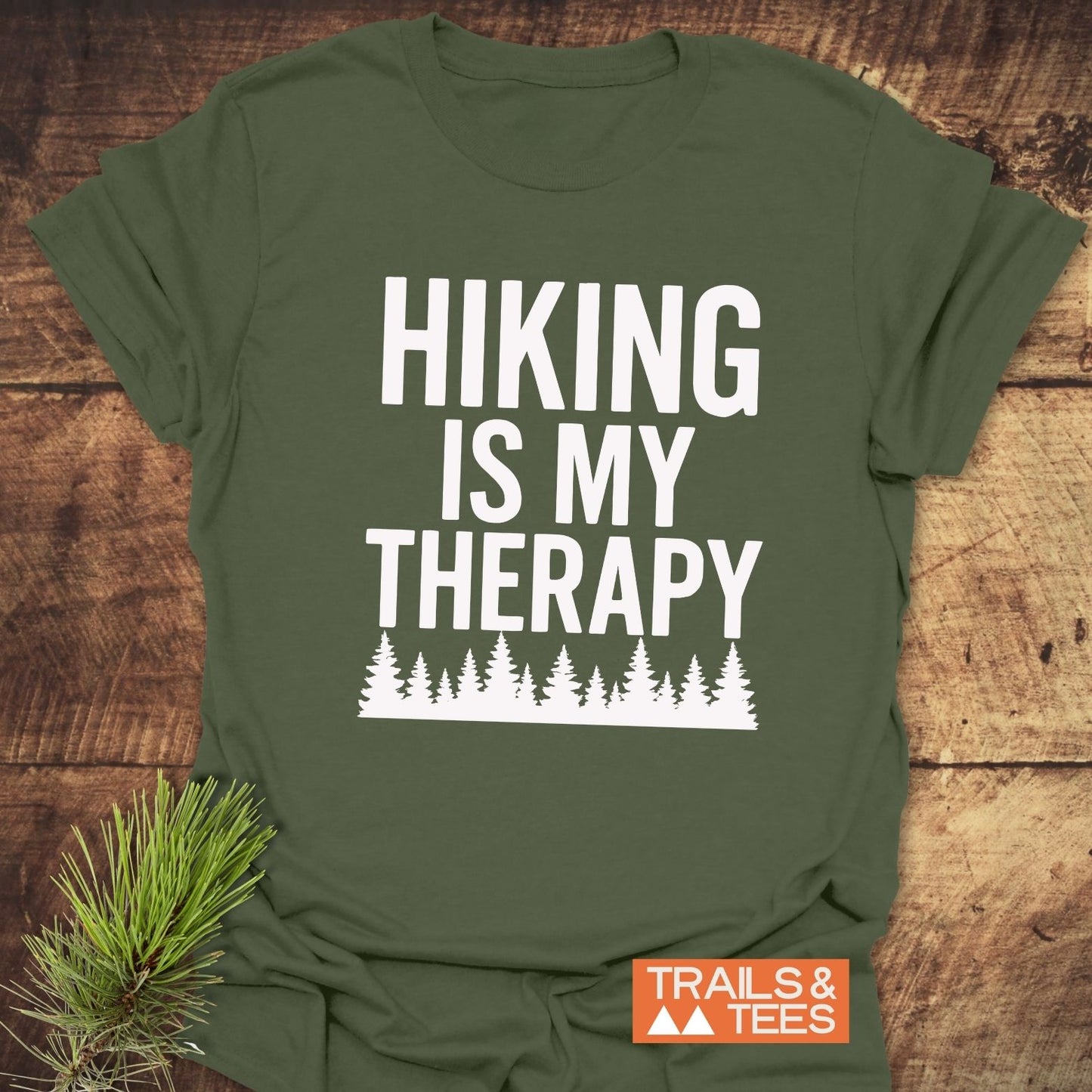 Hiking Is My Therapy T-Shirt on wood, featuring white slogan and pine trees. Soft, durable cotton; ideal for outdoor enthusiasts. Includes TRAILS & TEES label.