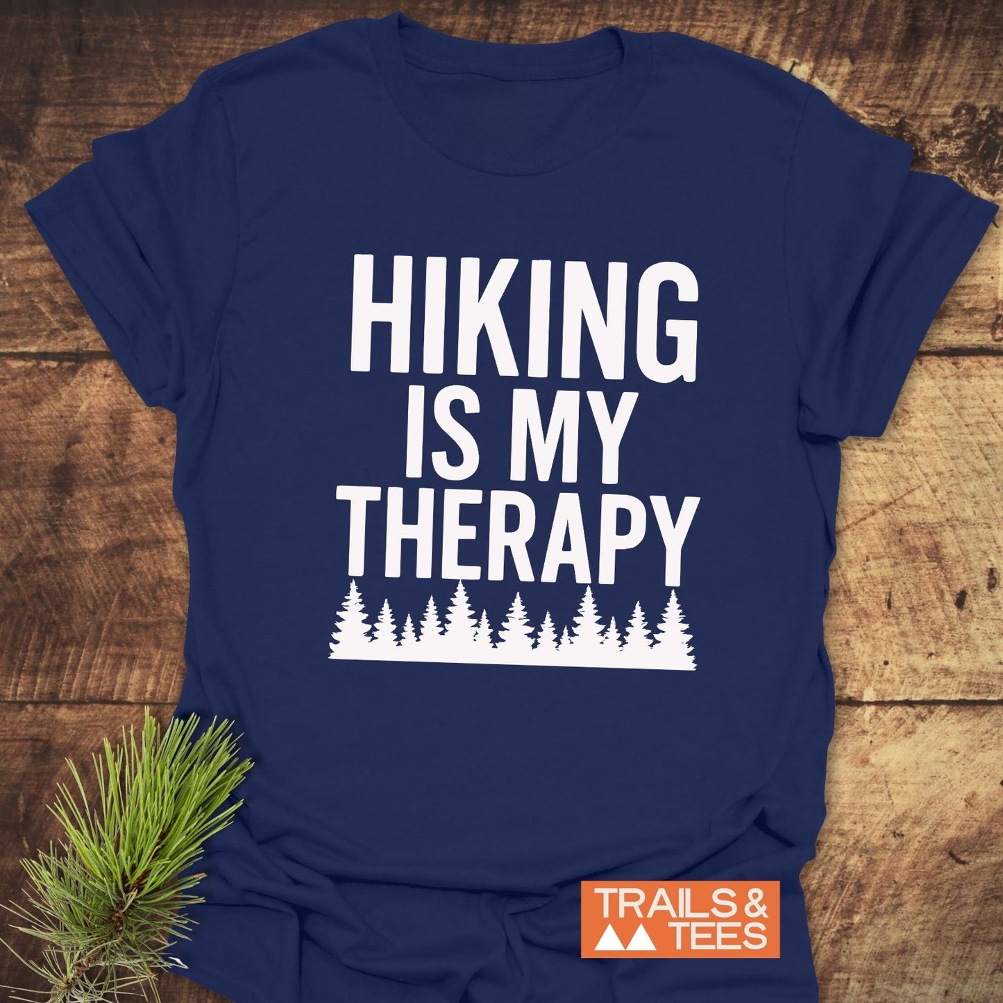Navy-blue Hiking Is My Therapy T-shirt with white slogan and pine-tree silhouettes, accented by a pine branch. Displays TRAILS & TEES logo.