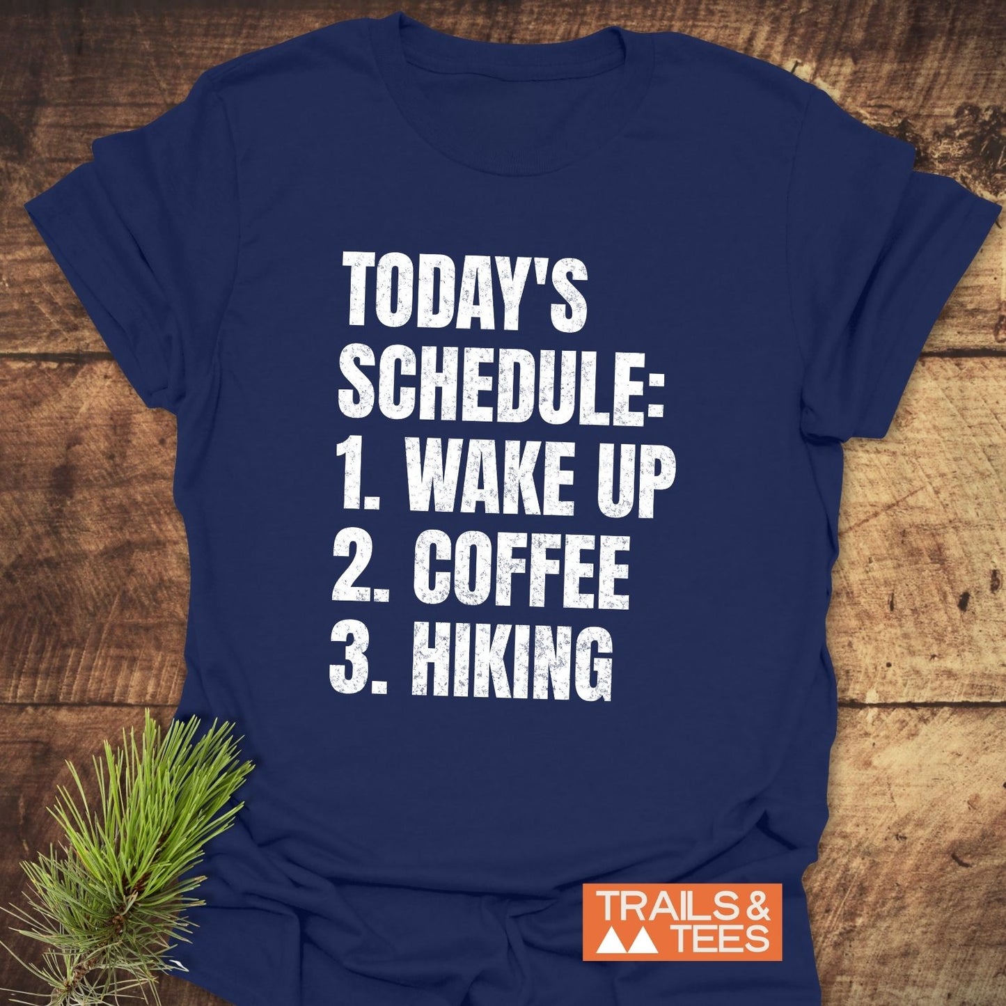 Today's Schedule Hiking T-Shirt on wood surface, featuring bold white lettering. Includes a small Trails & Tees logo, emphasizing comfort and outdoor adventure.