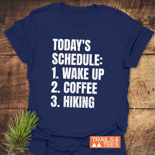 Today's Schedule Hiking T-Shirt on wood surface, featuring bold white lettering. Includes a small Trails & Tees logo, emphasizing comfort and outdoor adventure.