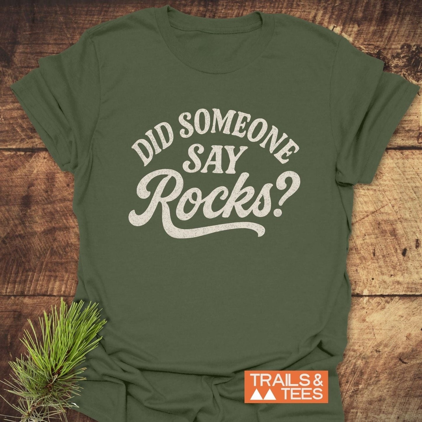 Did Someone Say Rocks? T-Shirt displayed on wood, featuring off-white text and TRAILS & TEES logo, with a pine sprig accentuating the outdoor theme.