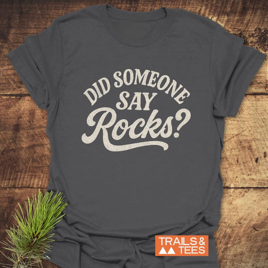 Did Someone Say Rocks? T-Shirt on wood, featuring distressed white script and Trails & Tees logo, with pine boughs. Soft cotton, durable print, pre-shrunk fit.