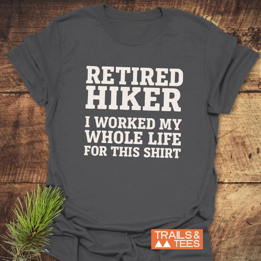 Dark gray Retired Hiker T-shirt with white text, orange TRAILS & TEES logo, on wood surface with pine sprig, highlighting outdoor adventures and comfort.