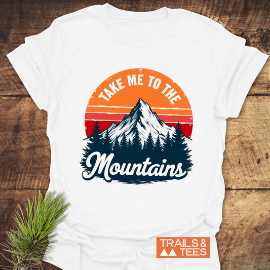 Take Me To The Mountains T-Shirt featuring a mountain and sunset print, crafted from soft cotton with durable, long-lasting design, laid flat with pine sprig accent.