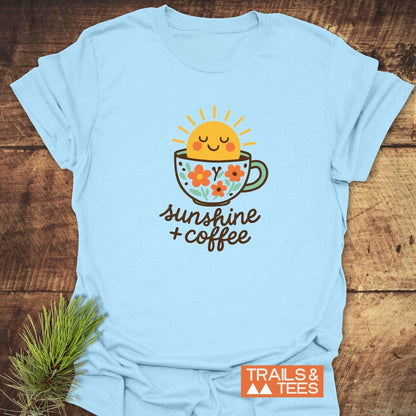 Sunshine And Coffee T-Shirt featuring a cute sun graphic in a floral coffee cup, hand-lettered text, and TRAILS & TEES logo on light-blue fabric.