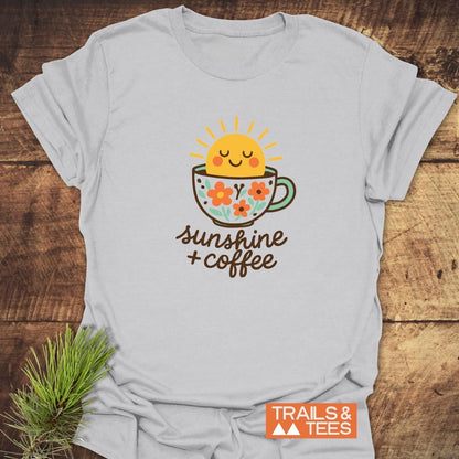 Sunshine And Coffee T-Shirt featuring a smiling sun in a coffee cup graphic and hand-lettered text, laid out on wood beside a pine sprig.