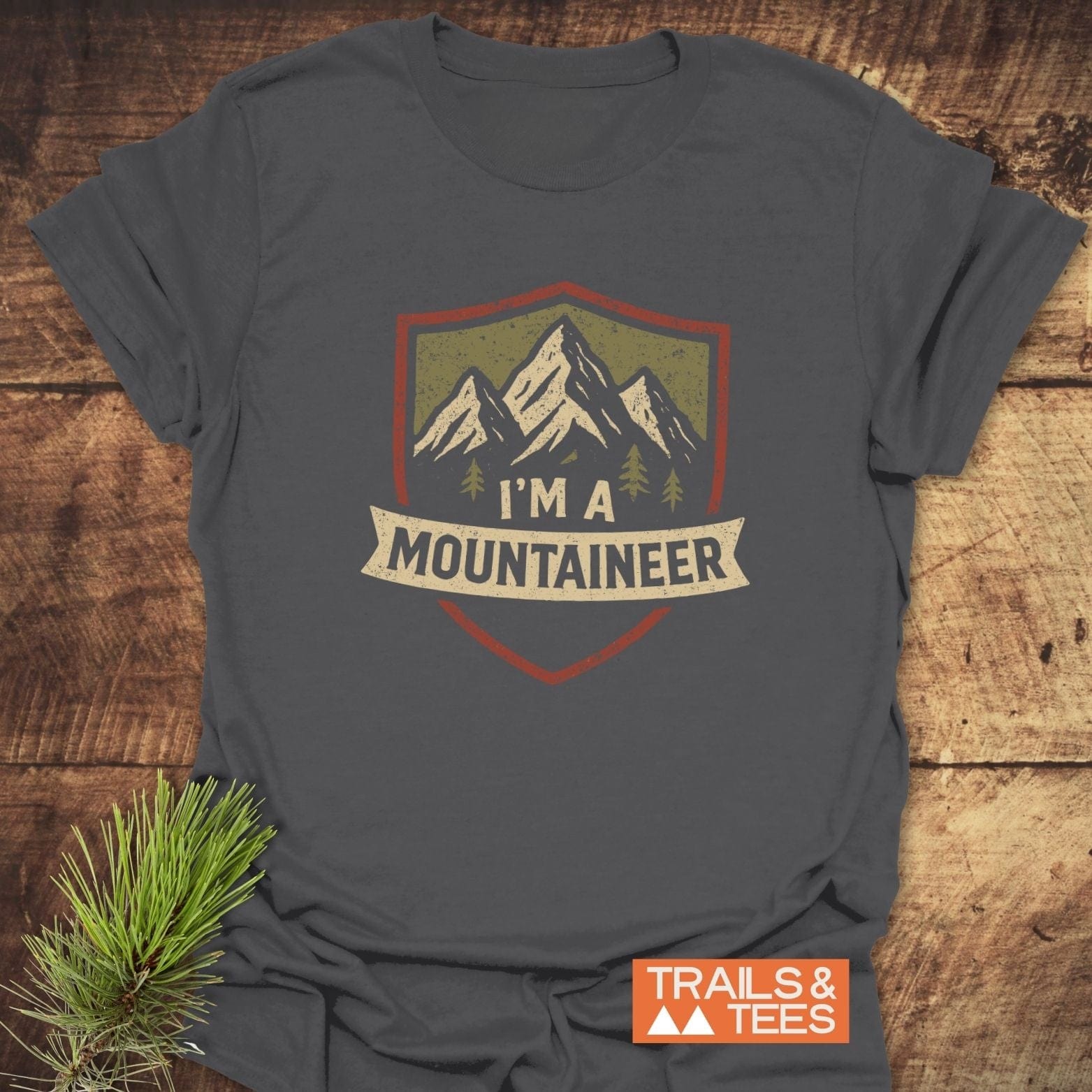 Mountaineer T-Shirt on rustic wood, featuring cream and olive mountain graphic with I'M A MOUNTAINEER banner. Includes TRAILS & TEES label and pine sprig.