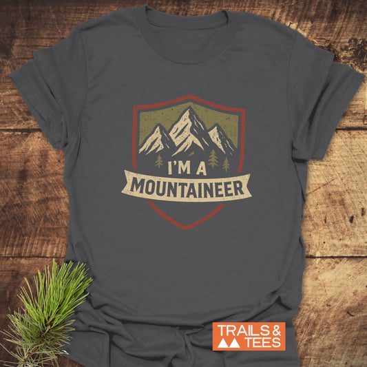 Mountaineer T-Shirt on rustic wood, featuring cream and olive mountain graphic with I'M A MOUNTAINEER banner. Includes TRAILS & TEES label and pine sprig.