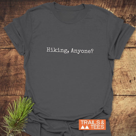 Dark gray Hiking, Anyone? T-shirt on wood, featuring a typewriter font and a TRAILS & TEES logo, complemented by pine needles.