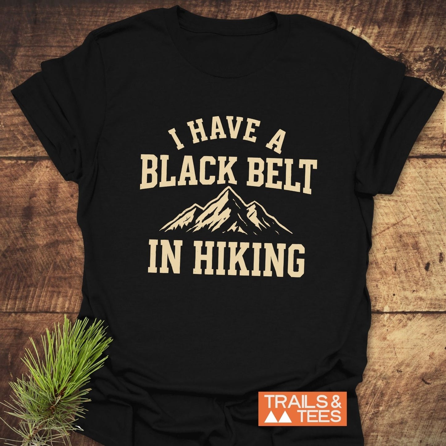 Black Belt In Hiking T-Shirt featuring a mountain graphic and quote, on wooden surface with TRAILS & TEES logo, emphasizing outdoor adventure spirit.
