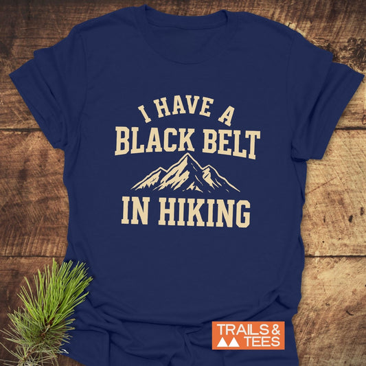 Navy Blue T-shirt with I HAVE A BLACK BELT IN HIKING in cream, featuring mountain peaks graphic. Trails & Tees logo in orange. Rustic wooden backdrop.