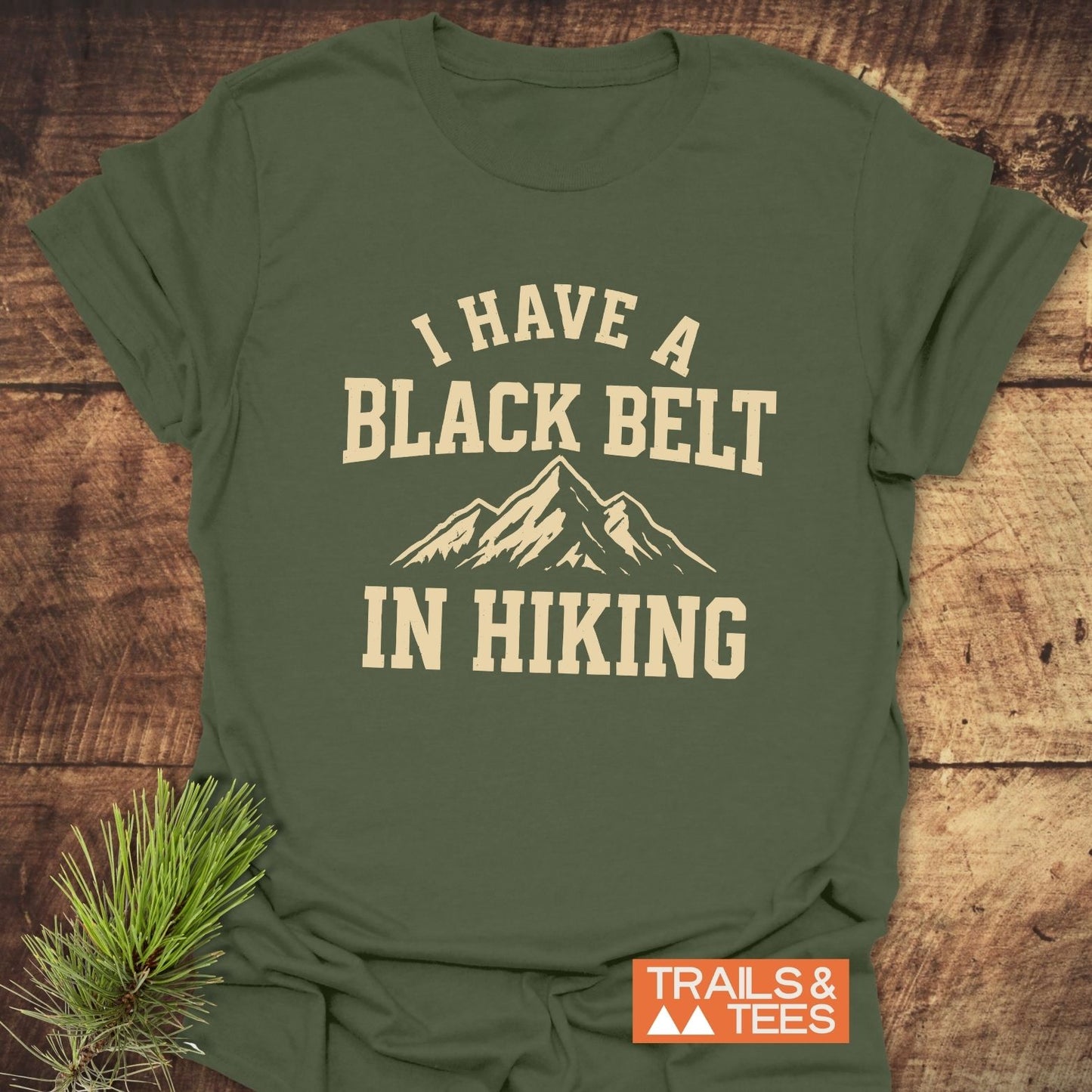 Olive-green T-shirt with I HAVE A BLACK BELT IN HIKING print, featuring a mountain graphic. Includes TRAILS & TEES logo and pine branch for styling.