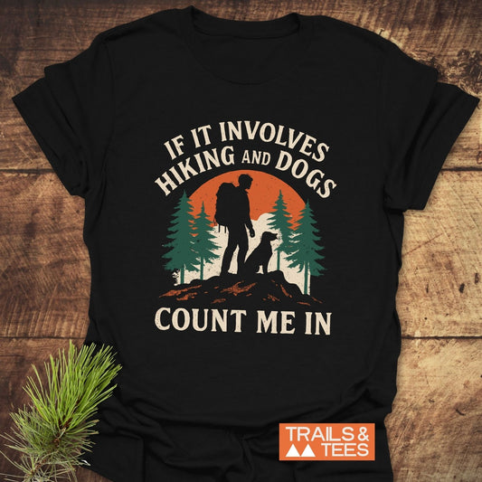 Hiking And Dogs T-Shirt with hiker and dog silhouette against sunset, slogan, and TRAILS & TEES logo on a wooden surface with pine sprig.