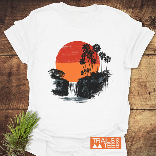 Sunset Waterfalls T-Shirt with a red-orange sun, palm trees, and waterfall design on soft cotton, laid out on wood with TRAILS & TEES logo.