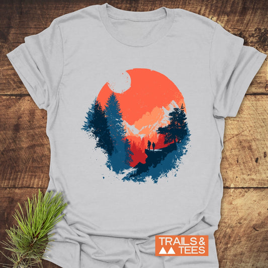 Sunset Hiking T-Shirt features a circular outdoor scene with an orange-red sun, snow-capped peaks, pine trees, and hikers; laid on rustic wood.