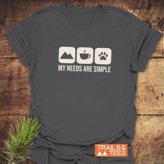 My Needs Are Simple T-Shirt featuring mountain, coffee, and paw print icons, with 'TRAILS & TEES' logo, styled with evergreen sprig on wooden surface.