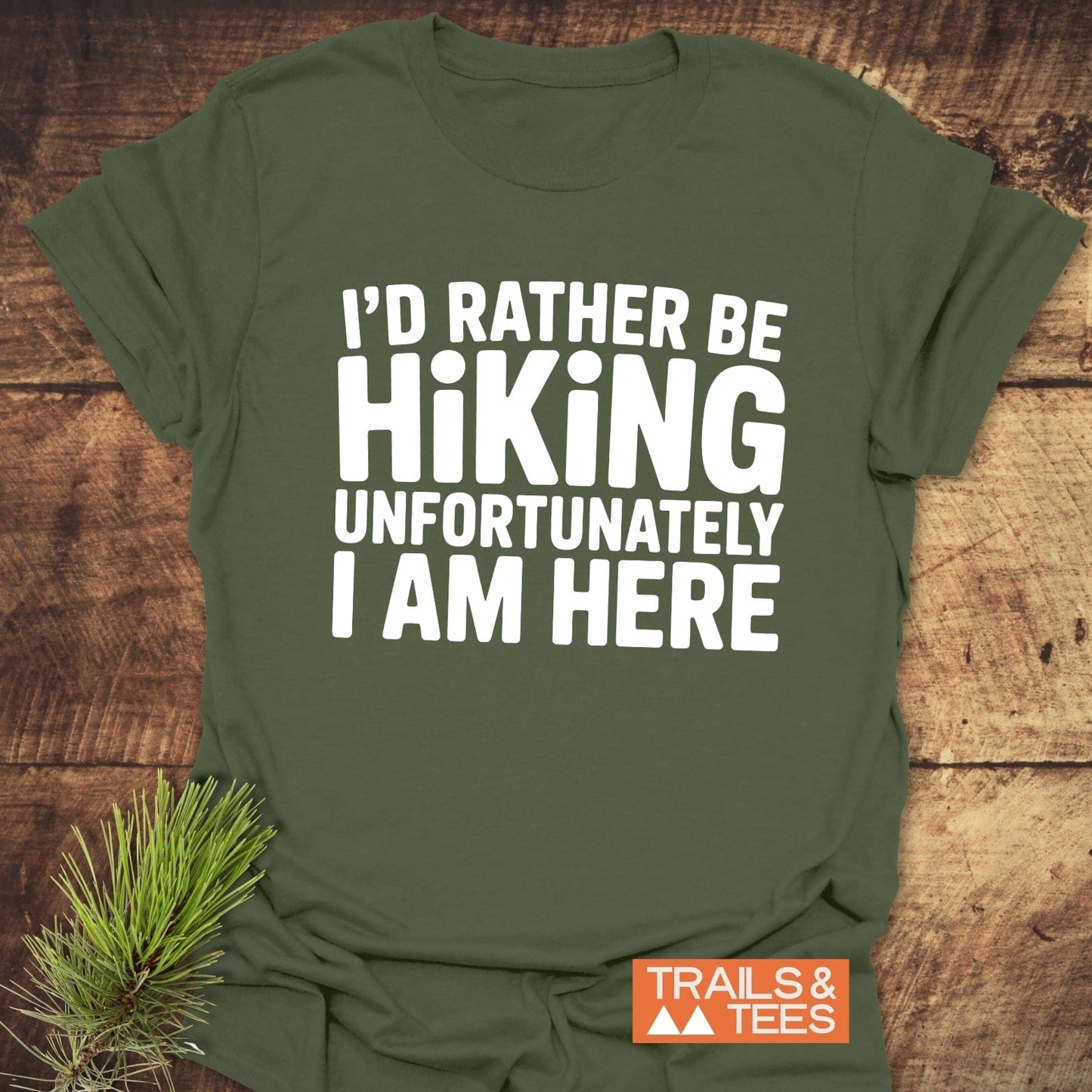 I'd Rather Be Hiking... T-Shirt displayed on wood, featuring bold white slogan, emphasizing outdoor theme with pine needles, crafted from soft cotton for comfort and durability.