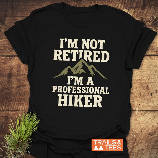 I'm Not Retired T-Shirt featuring an olive-green mountain silhouette and slogan, laid on wood with pine sprig. Logo: TRAILS & TEES.