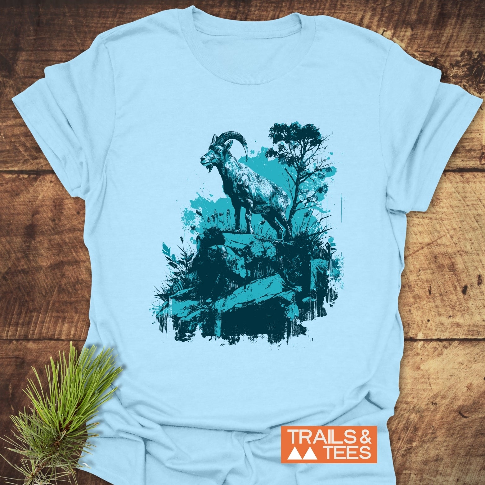 Wild and Steady – Mountain Goat T-Shirt featuring a stylized goat print, crafted from soft cotton, on a wooden surface with a pine sprig.