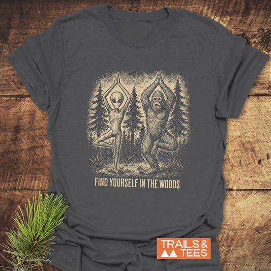 Find Yourself In The Woods T-Shirt: Flat-lay of charcoal tee featuring alien and Bigfoot in yoga pose, captioned for outdoor enthusiasts. Made from soft, durable, ring-spun cotton.