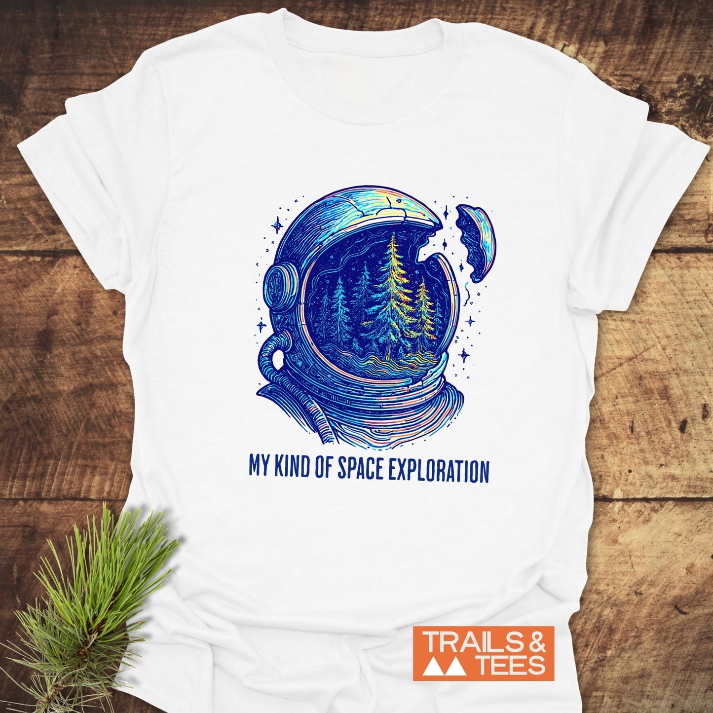 My Kind Of Space Exploration T-Shirt featuring a cracked astronaut helmet revealing pine trees, placed on wood with a pine sprig, logo TRAILS & TEES visible.