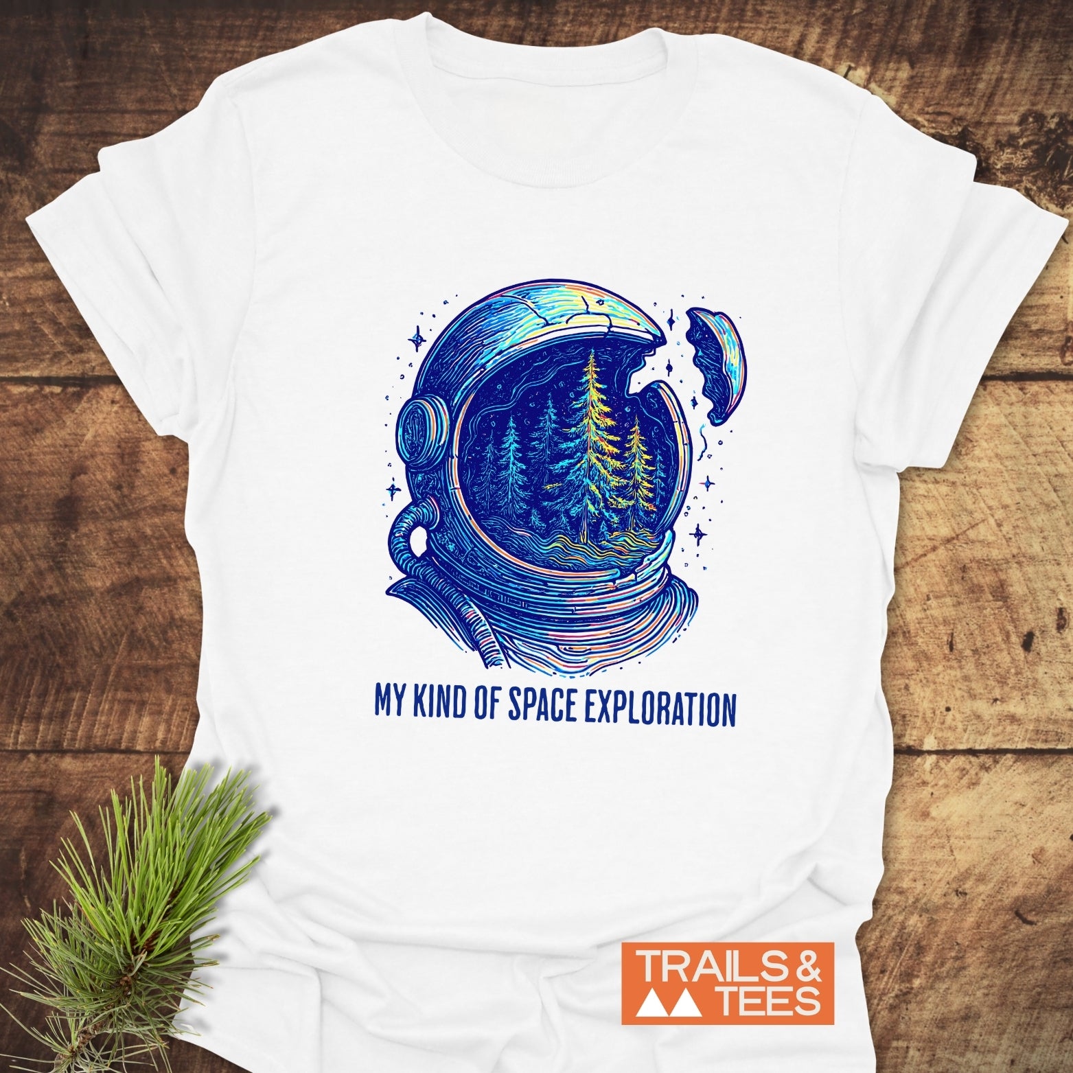 My Kind Of Space Exploration T-Shirt featuring a cracked astronaut helmet revealing pine trees, placed on wood with a pine sprig, logo TRAILS & TEES visible.