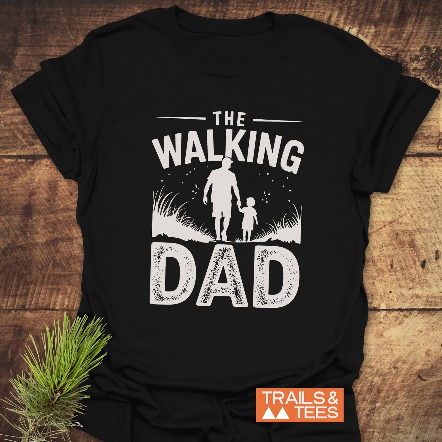 The Walking Dad T-Shirt on wooden surface, featuring white text and dad-child silhouette graphic; includes 'TRAILS & TEES' logo, crafted for comfort and outdoor enthusiasts.