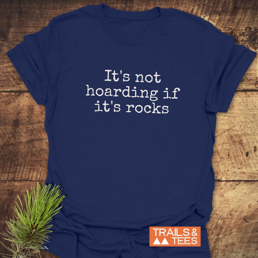 Navy-blue T-shirt with It’s not hoarding if it’s rocks slogan, typewriter-style font, small orange Trails & Tees logo, laid on wooden surface with pine sprig.