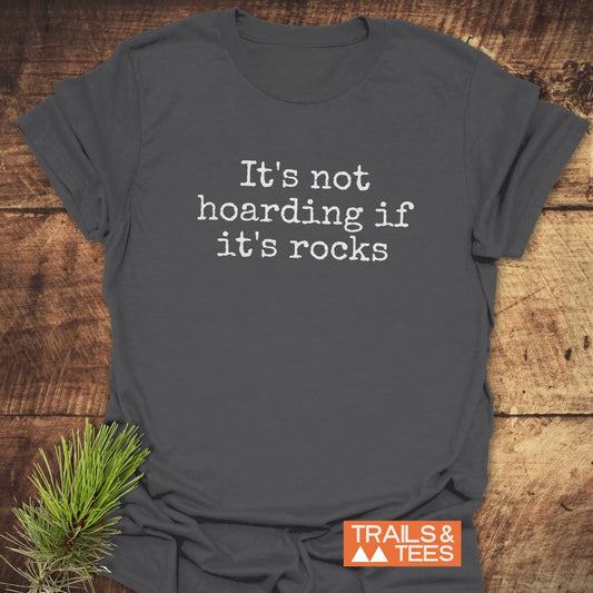 Dark gray It's Not Hoarding If It's Rocks T-shirt on wood surface, featuring white typewriter-style text and TRAILS & TEES logo, with pine greenery accent.
