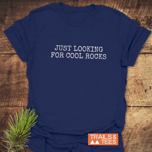 Navy-blue Just Looking For Cool Rocks T-shirt with typewriter-style lettering, laid on wood. Features Trails & Tees logo. Soft, durable, outdoor-themed design.