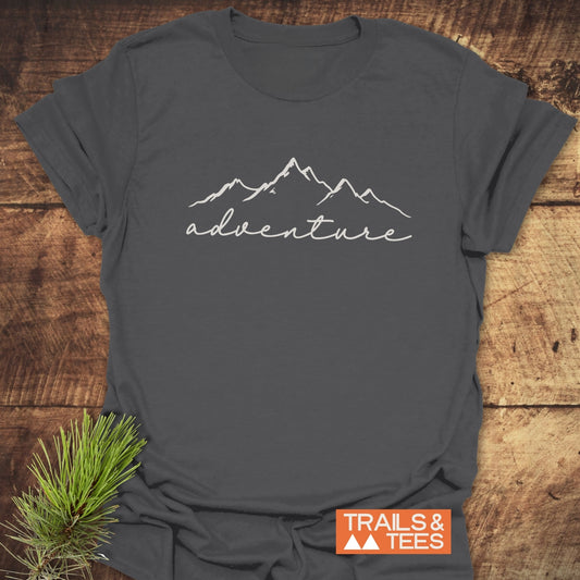 Heather-charcoal Adventure T-Shirt with mountain ridge design, laid on wood. Features durable print, soft cotton, pre-shrunk for fit. Outdoorsy touch with pine sprig.
