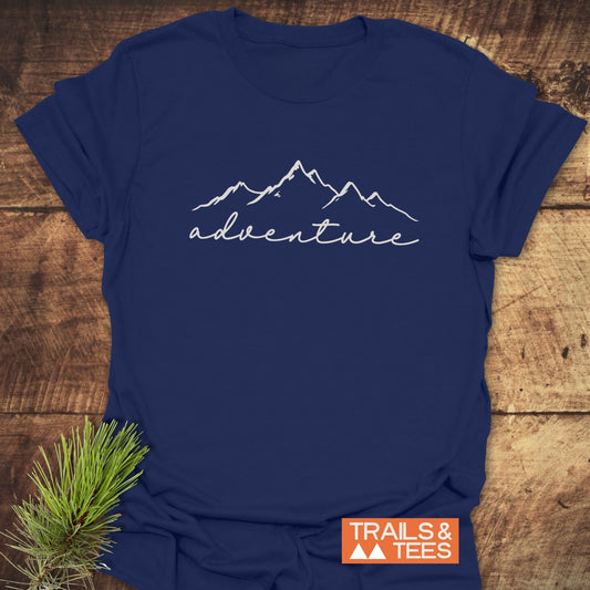 Adventure T-Shirt featuring a mountain range line-drawing and “adventure” text, laid flat with rustic elements. Crafted from soft cotton for outdoor enthusiasts.