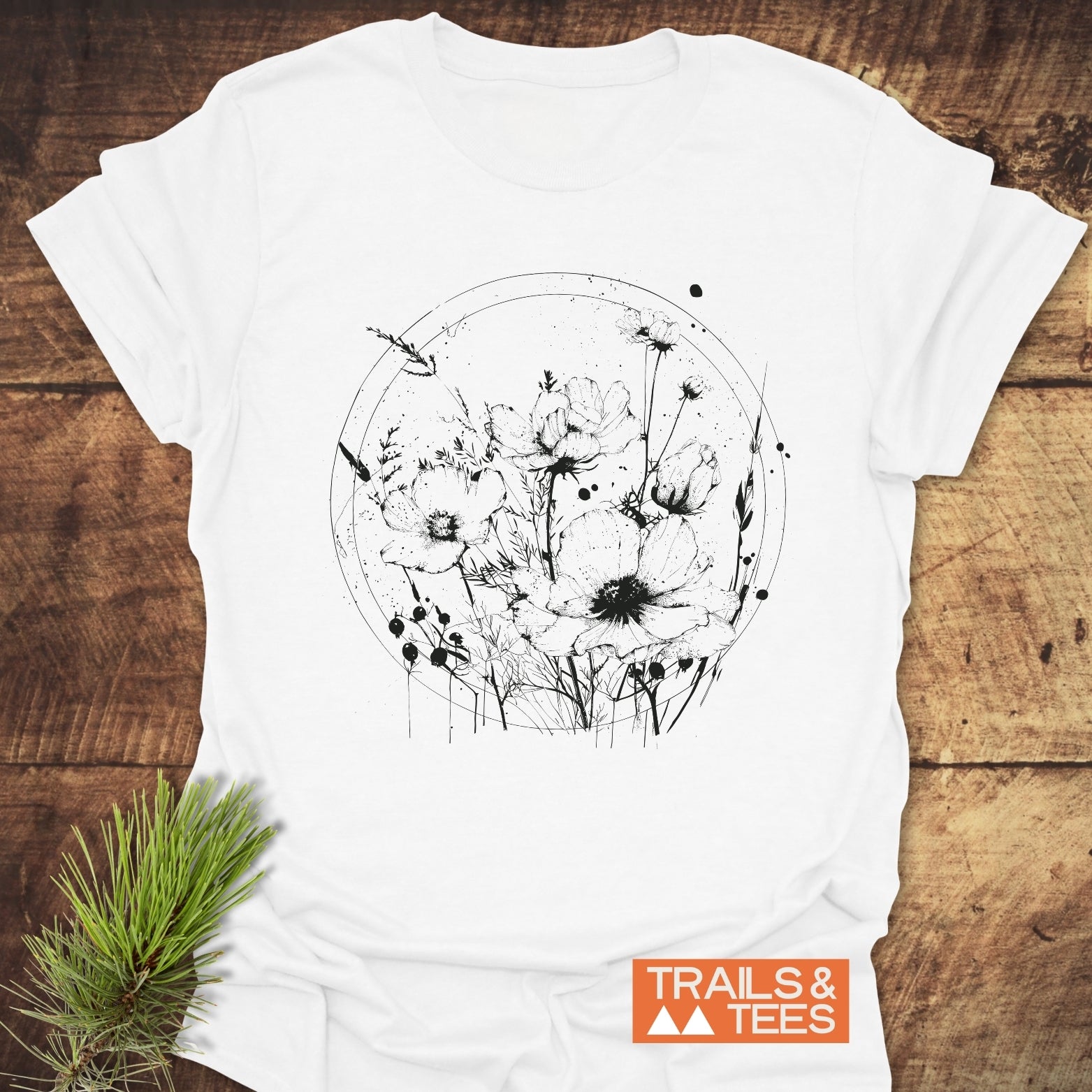 Wildflower Circle T-Shirt featuring black line-art of poppies and grasses, styled with evergreen sprig, showcasing Trails & Tees logo on rustic wooden surface.