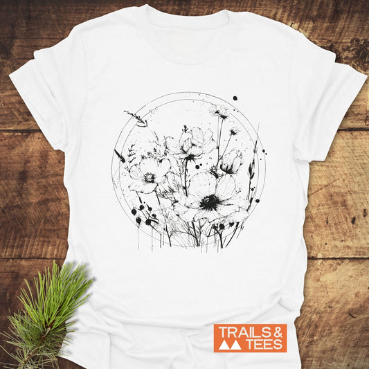 Wildflower Circle T-Shirt featuring black line-art of poppies and grasses, styled with evergreen sprig, showcasing Trails & Tees logo on rustic wooden surface.