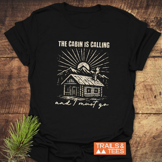 The Cabin Is Calling T-Shirt features a rustic log cabin design with mountain peaks and sun. Crafted from soft ring-spun cotton for adventure-ready comfort.