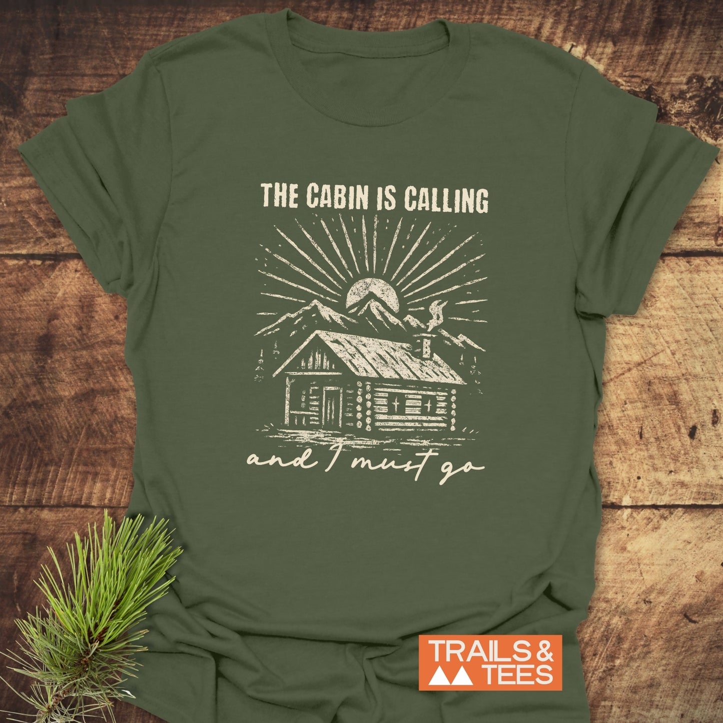 Olive-green T-shirt with THE CABIN IS CALLING graphic of a log cabin, mountains, and sun, symbolizing outdoor adventure. Includes a pine sprig and TRAILS & TEES logo.
