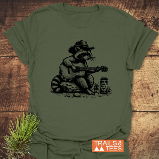 Guitar Playing Raccoon T-Shirt features a raccoon in a cowboy hat playing guitar, screen-printed on a soft, olive-green tee, embodying outdoor adventure spirit.