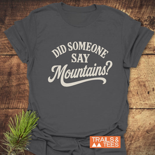 Charcoal-gray “Did Someone Say Mountains?” T-shirt with off-white print, laid flat. Features a small Trails & Tees logo and pine sprig beside it.