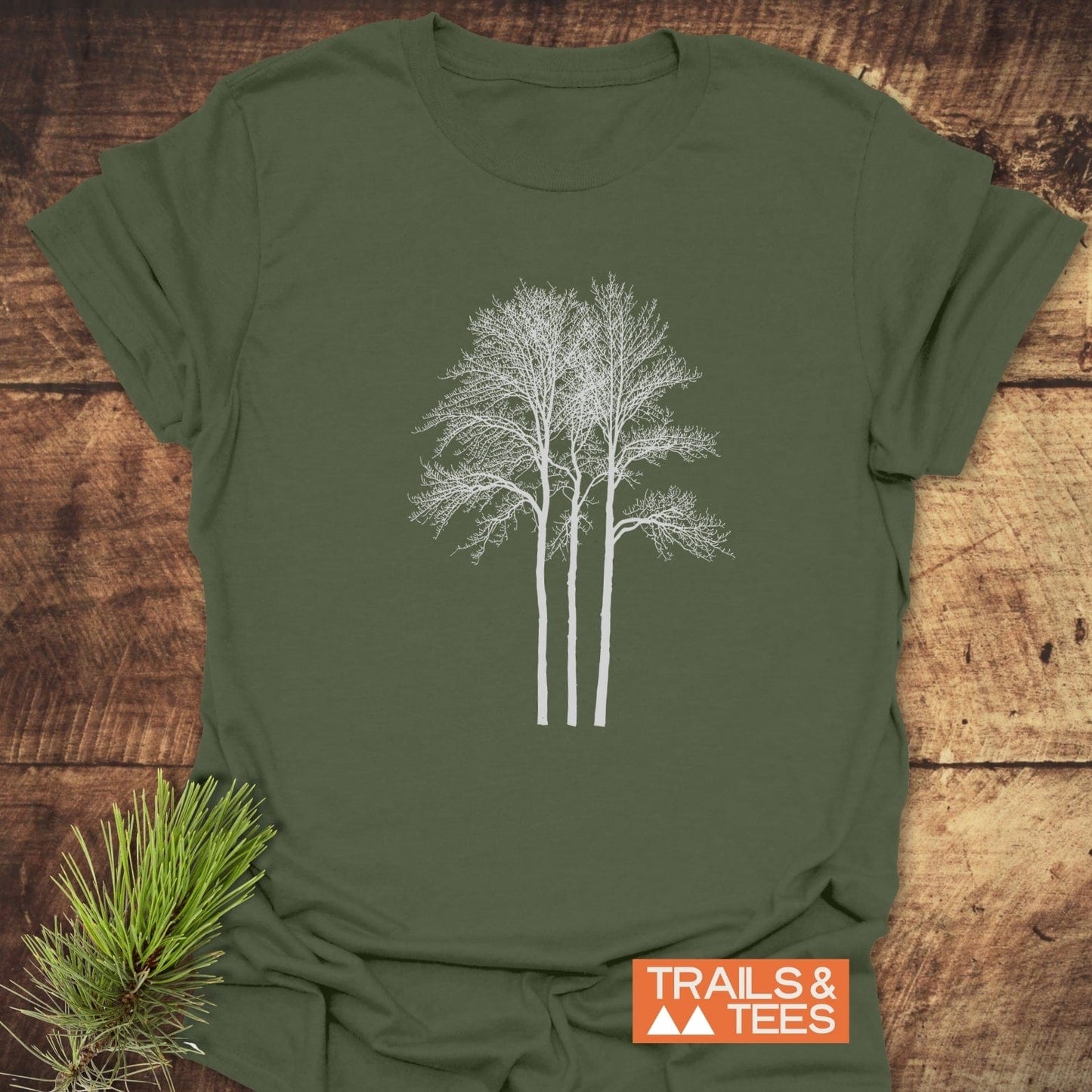 Tree Silhouettes T-Shirt on wood surface, featuring white tree graphic. Includes TRAILS & TEES logo and pine sprig, highlighting outdoor-inspired design and comfort.