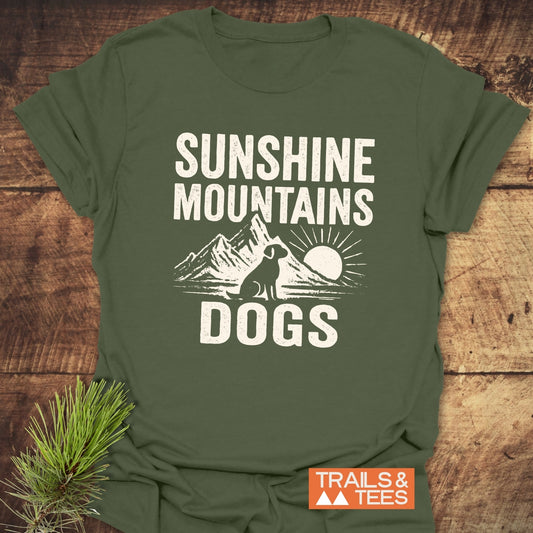 Sunshine Mountains Dogs T-Shirt on wood surface with pine sprig, featuring a mountain dog graphic and TRAILS & TEES label. Made from soft, durable cotton.