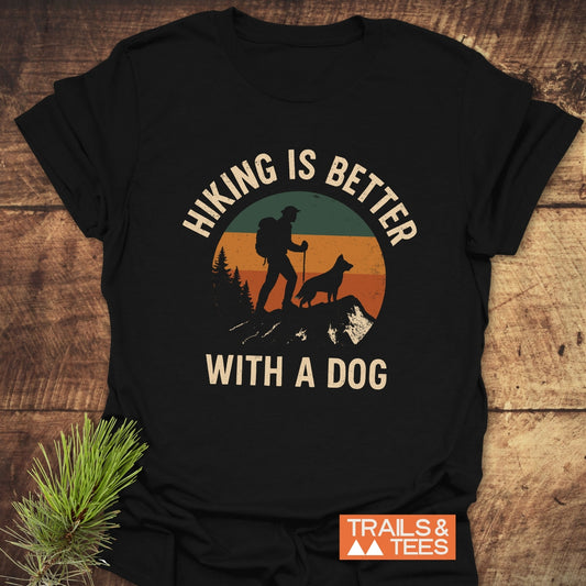 Hiking Is Better With A Dog T-Shirt featuring a retro hiker graphic, crafted from soft cotton for comfort, laid on a wooden surface.