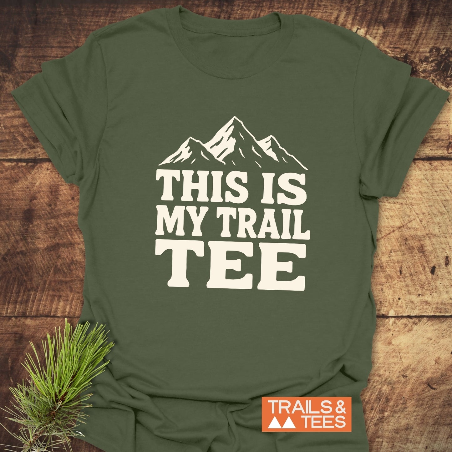 The This Is My Trail Tee T-Shirt features mountain graphics and text, displayed on a wooden surface with pine needles, embodying an outdoorsy, adventure-ready style.