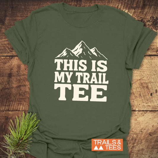 The This Is My Trail Tee T-Shirt features mountain graphics and text, displayed on a wooden surface with pine needles, embodying an outdoorsy, adventure-ready style.
