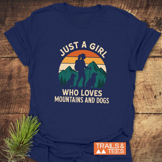 Mountains And Dogs T-Shirt featuring a hiker and dogs graphic, slogan, and TRAILS & TEES logo, laid on a wooden surface.