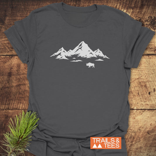 Mountains And Buffalo T-Shirt on rustic surface, featuring a white mountain and buffalo print. Includes TRAILS & TEES logo; pine needles accent the scene.
