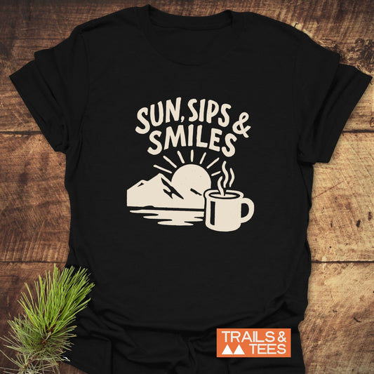 Sun, Sips, Smiles T-Shirt on rustic wood, featuring a sun, mountain, and mug graphic. Made from soft cotton, ideal for outdoor enthusiasts.