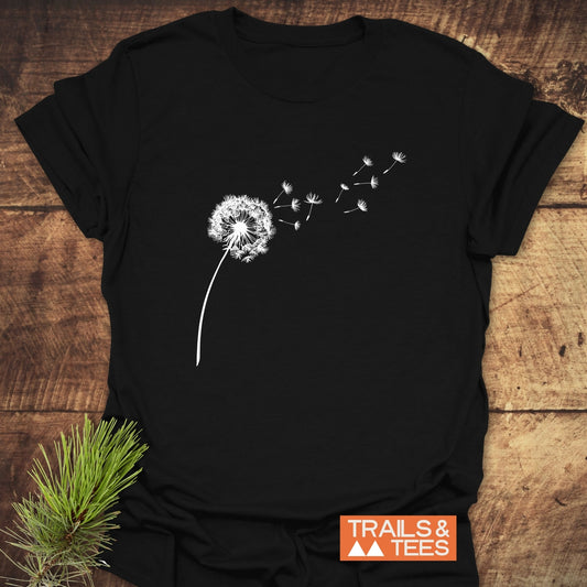 Dandelion T-Shirt: Black tee with white dandelion silhouette, featuring orange TRAILS & TEES logo, on wooden surface. Crafted for comfort and durability.