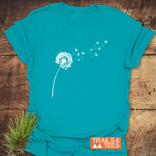 Dandelion T-Shirt featuring a white dandelion silhouette with seeds blowing right, laid on wood, emphasizing comfort and durability for outdoor enthusiasts.